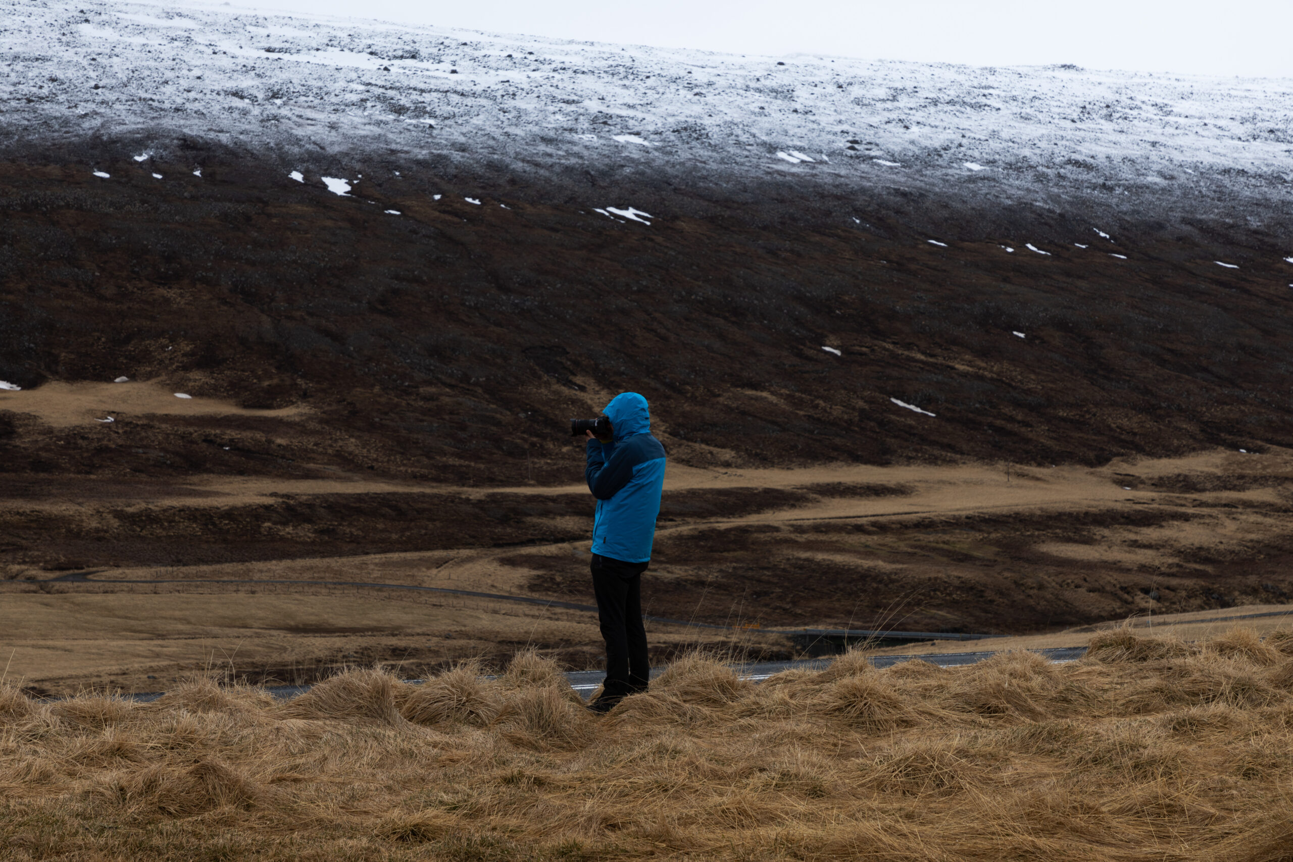 A photographer in a blue jacket captures Iceland’s rugged landscape on 15 May 2022. They stand on a grassy field, framed by a snow-dusted hillside under an overcast sky, highlighting the stark contrast between the earthy foreground and the mountainous backdrop.