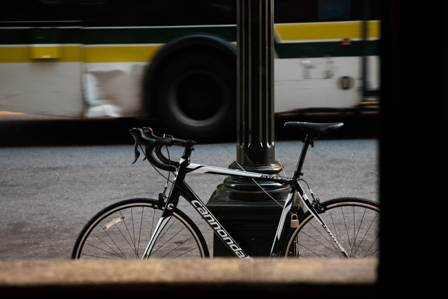 A sleek black and white Cannondale Synapse road bike is locked to a lamppost on an urban street, with a passing bus blurred in the background. The scene captures a moment of city life, contrasting the stillness of the bike with the motion of public transit.